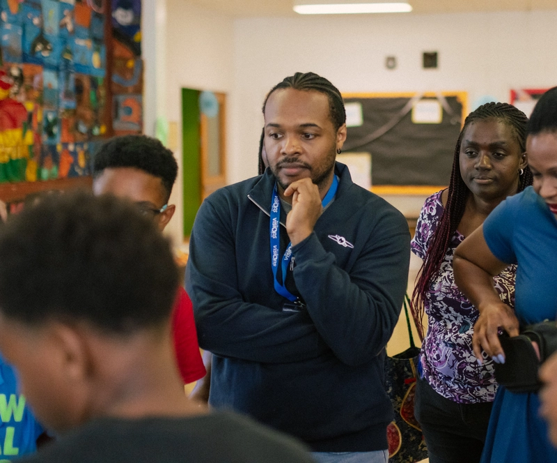 A group of adults and children gather closely, listening intently in a colorful classroom setting.