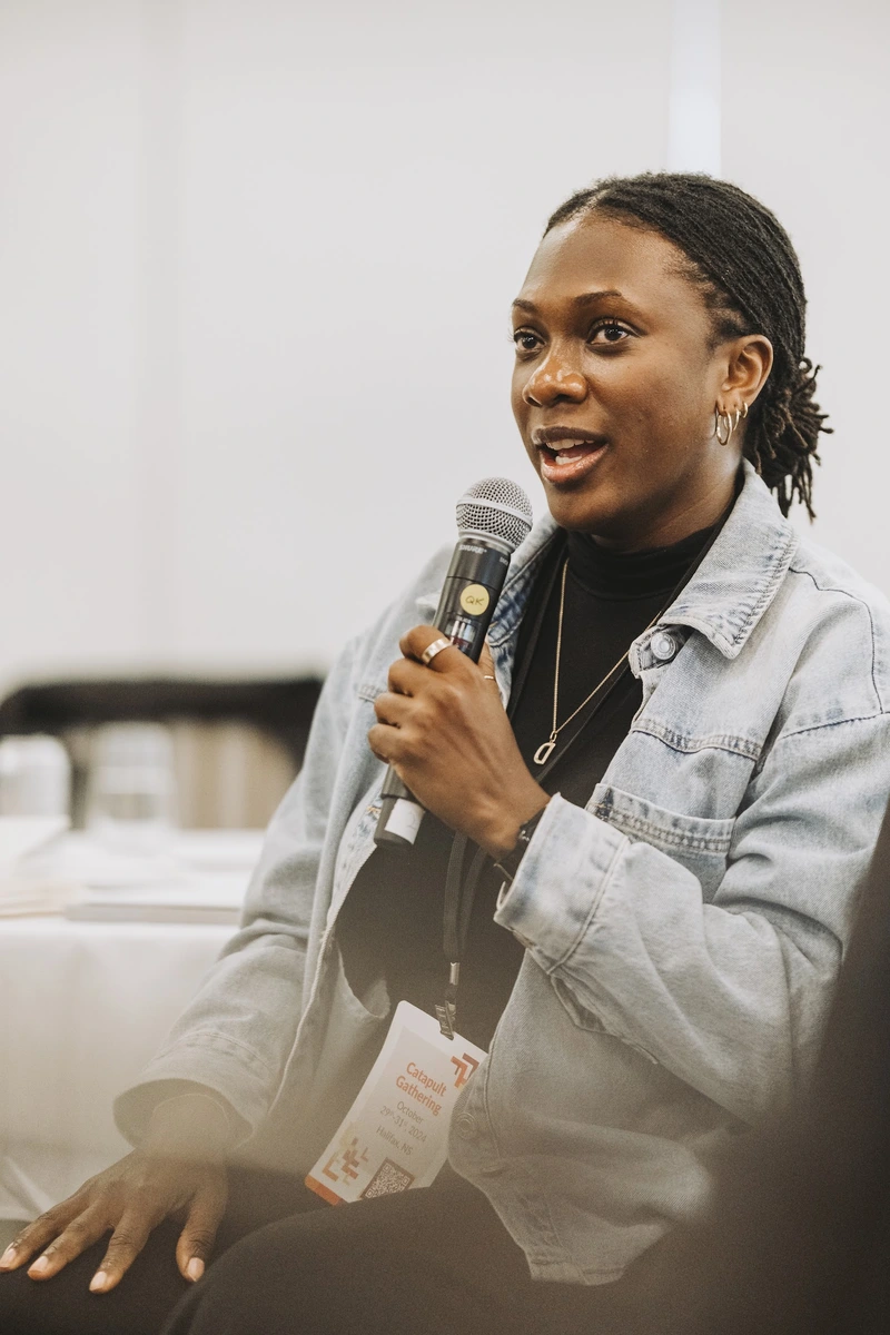 A woman speaks into a microphone while seated, wearing a denim jacket and a conference name badge.