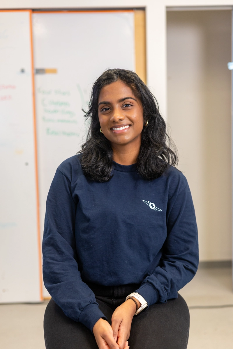 A woman with dark hair in a navy sweatshirt sits and smiles in a bright, indoor setting.