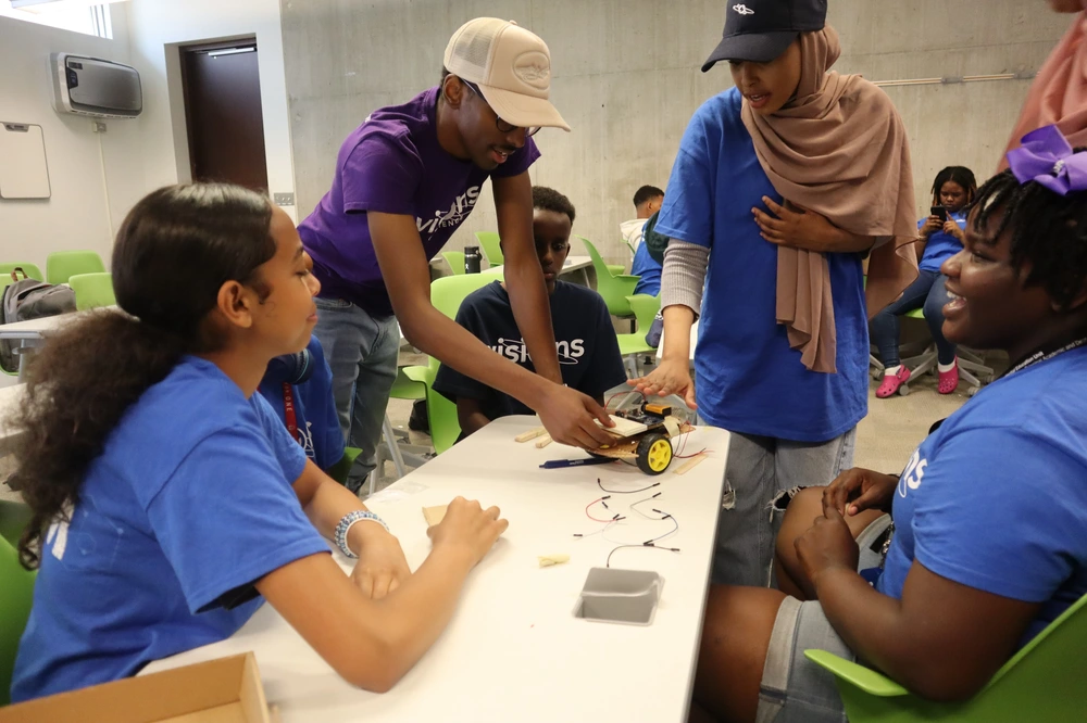 Students gather around a table working on a small wheeled robot in a classroom setting.