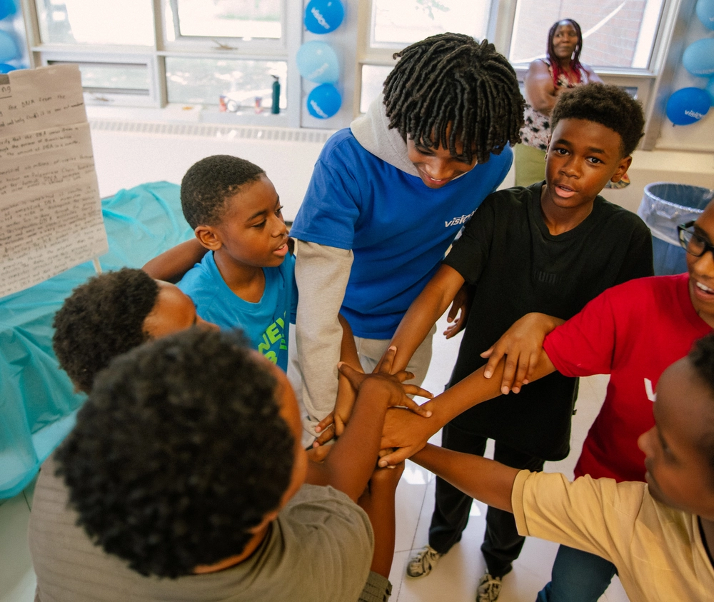 A group of children put their hands together in a team huddle inside a classroom decorated with blue balloons.