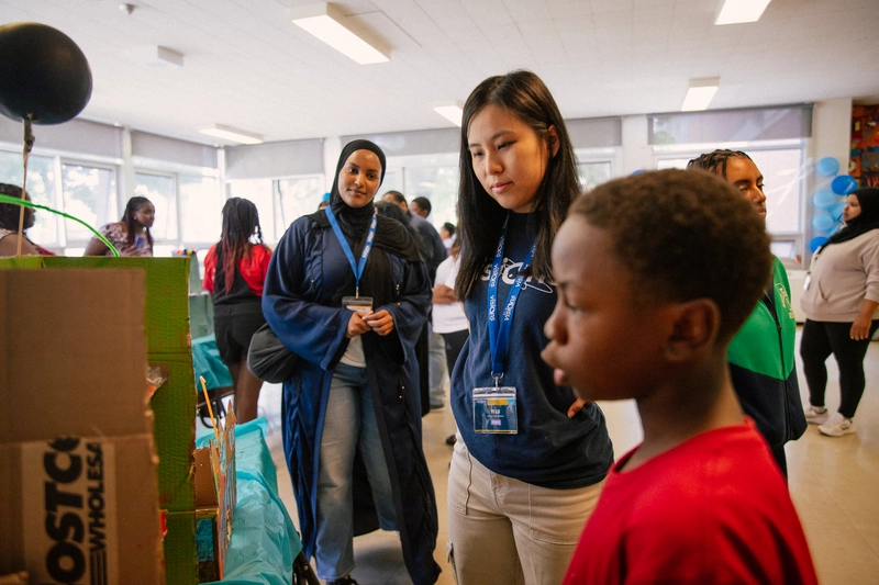 Children and adults gather indoors, observing displays and projects at a community event.