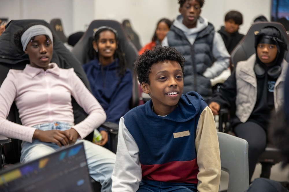 Several kids sit and watch something intently in a classroom or group setting, some smiling and engaged.