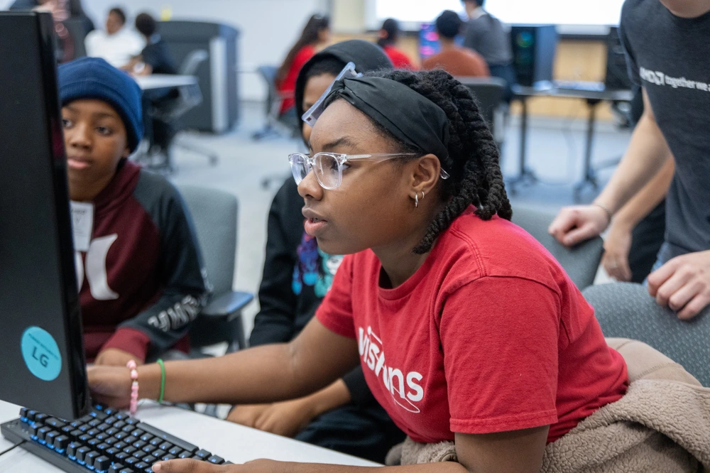 A girl in glasses and a red shirt focuses on a computer screen in a classroom with other students.
