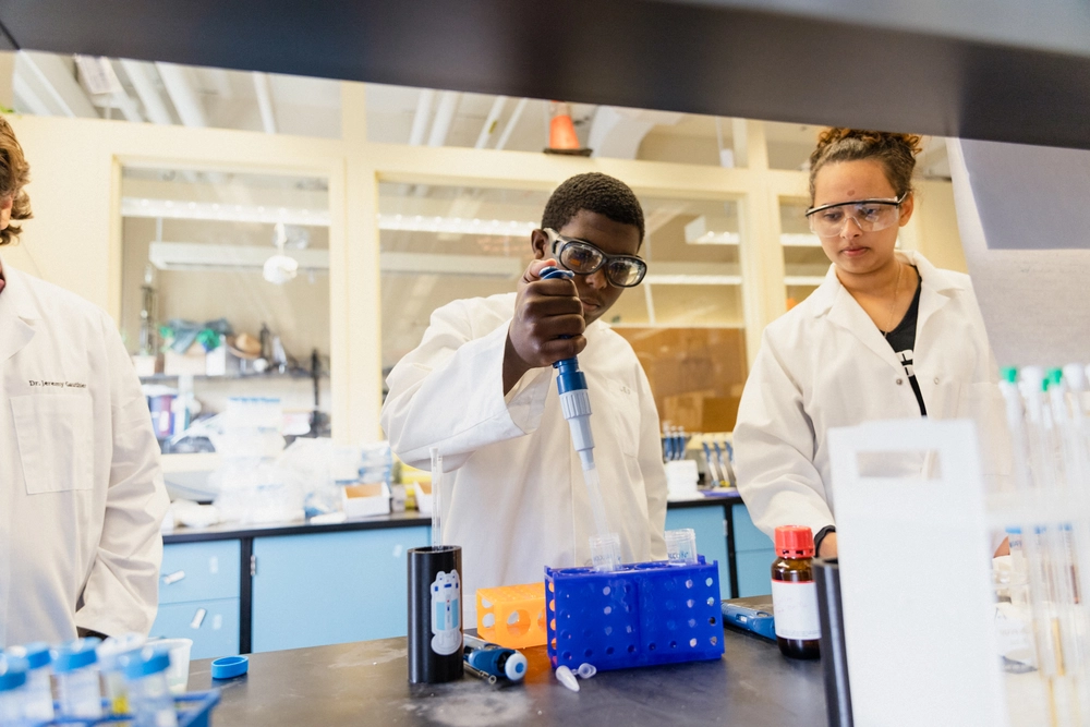 Two students in lab coats and goggles conduct an experiment with test tubes and a pipette in a science lab.