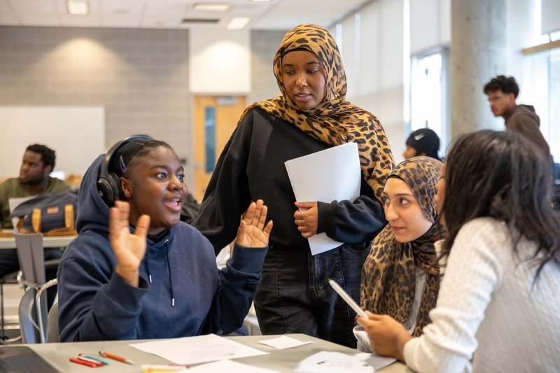 Four students sit and stand around a table, talking and working together in a bright classroom.