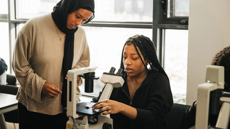 Two students work together using a microscope in a classroom with large windows in the background.