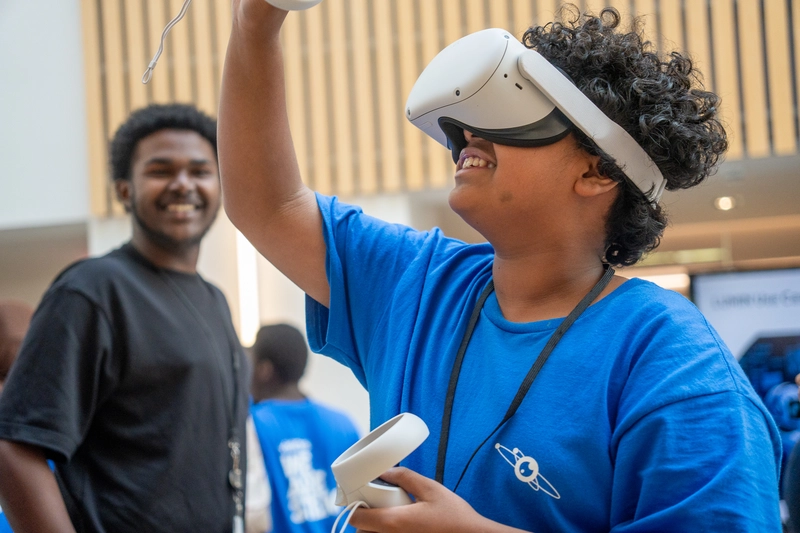 A smiling boy in a blue shirt uses a VR headset, while another boy smiles in the background.