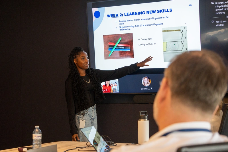 A woman presents a slide titled Week 2: Learning New Skills to a group in a conference room.