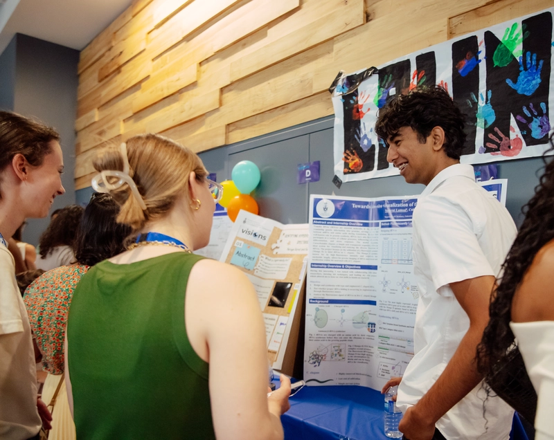 Three people view a science fair poster while talking to its presenter in a bright, decorated hallway.