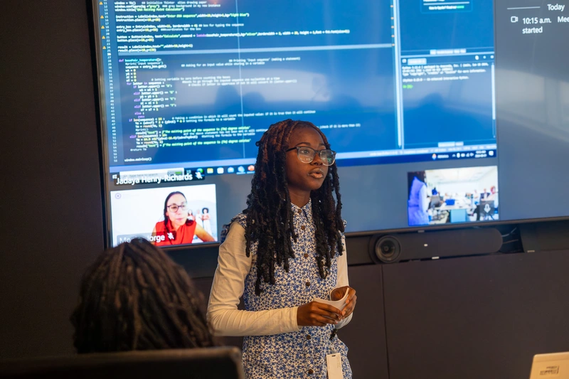Young woman presents in front of a large screen displaying code and a virtual meeting during a tech session.