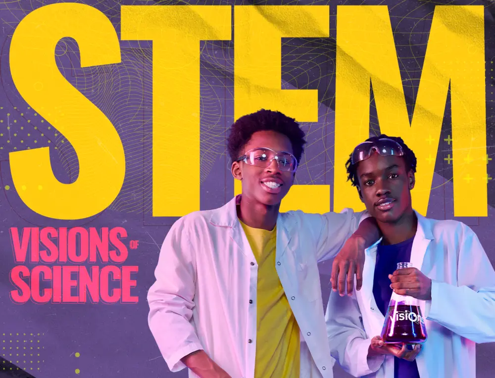 Two young men in lab coats and goggles stand smiling in front of a large STEM sign, holding a beaker.