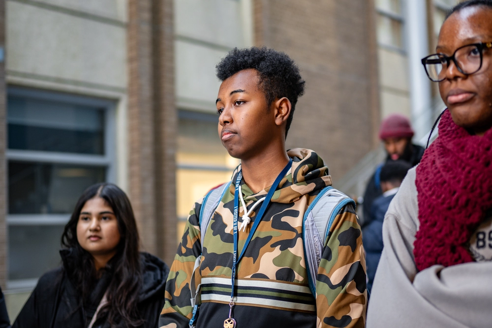 Three students stand outdoors, one wearing a camo hoodie, looking thoughtful, with a building in the background.