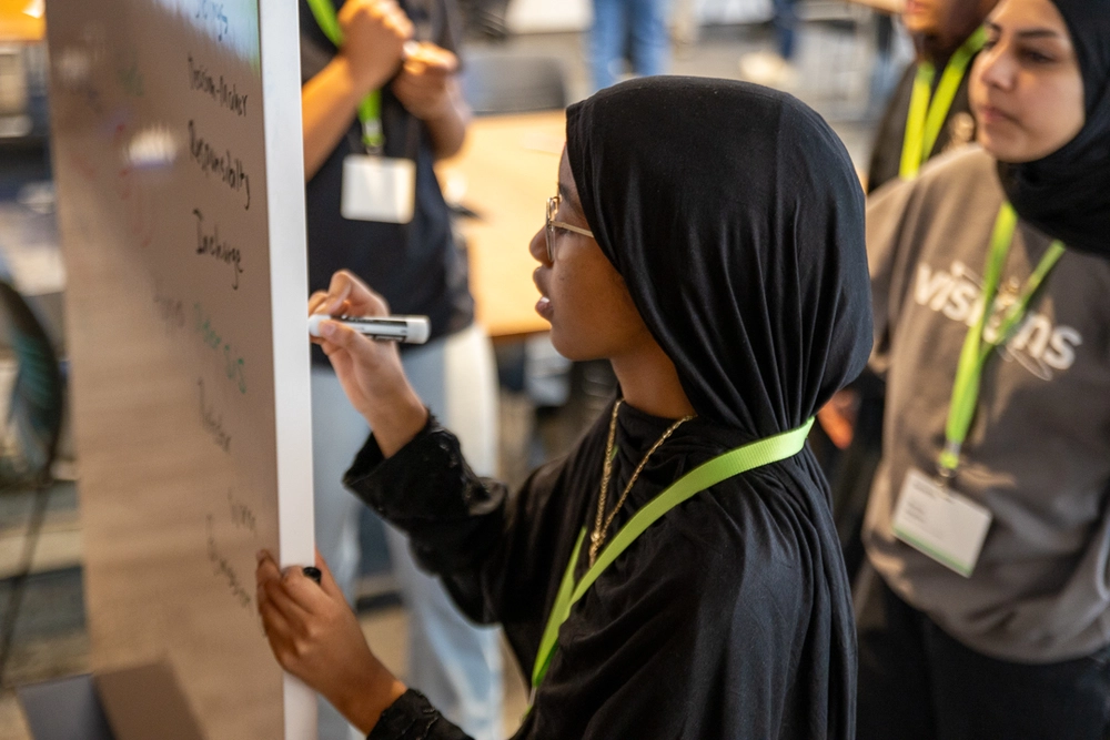A woman in a black hijab writes on a whiteboard while others watch in a classroom setting.