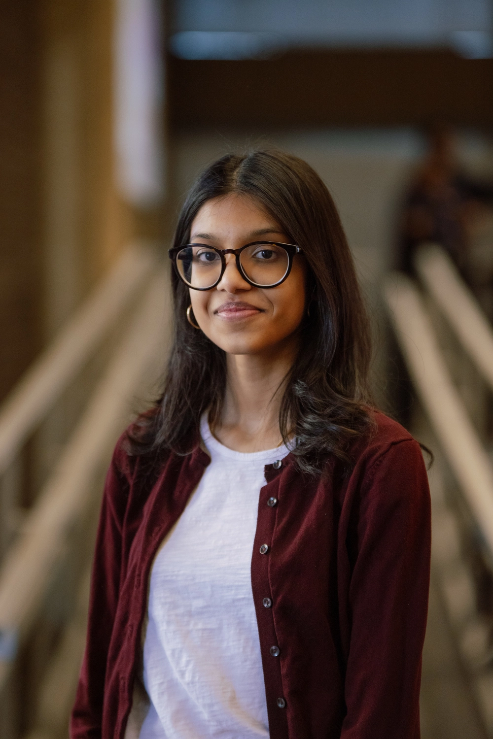 Young woman with long dark hair and glasses, wearing a white shirt and maroon cardigan, smiling slightly.