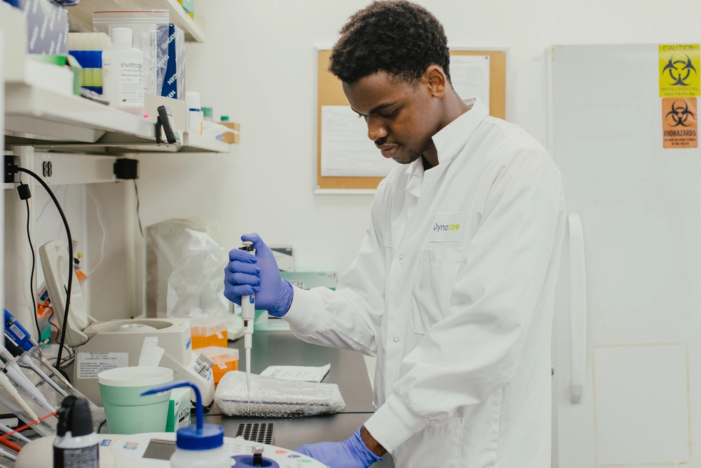 A scientist in a lab coat uses a pipette at a laboratory workstation with equipment and supplies nearby.