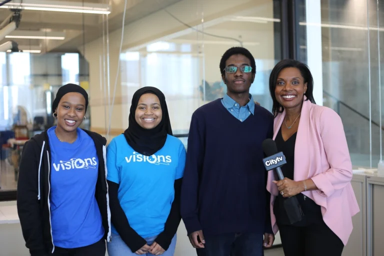 Four people smile in an office; two wear blue Visions shirts and one holds a Citytv microphone.