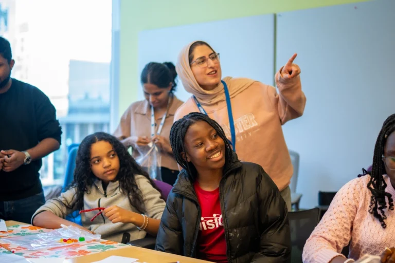 A group of young people work on crafts at a table while one woman smiles and points to something off-camera.
