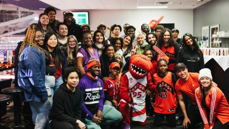 Group of people posing and smiling with the Toronto Raptors basketball mascot indoors, wearing team gear.
