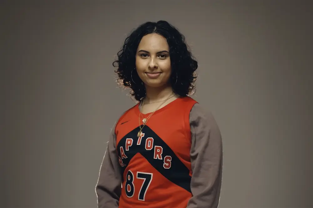 Person with curly hair wearing a red Raptors jersey and gray sleeves, smiling at the camera against a plain background.