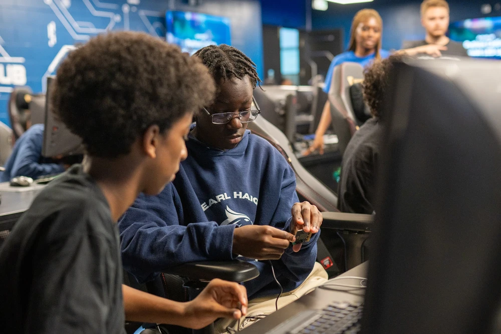 Two students work together with electronics in a computer lab, while others are visible in the background.
