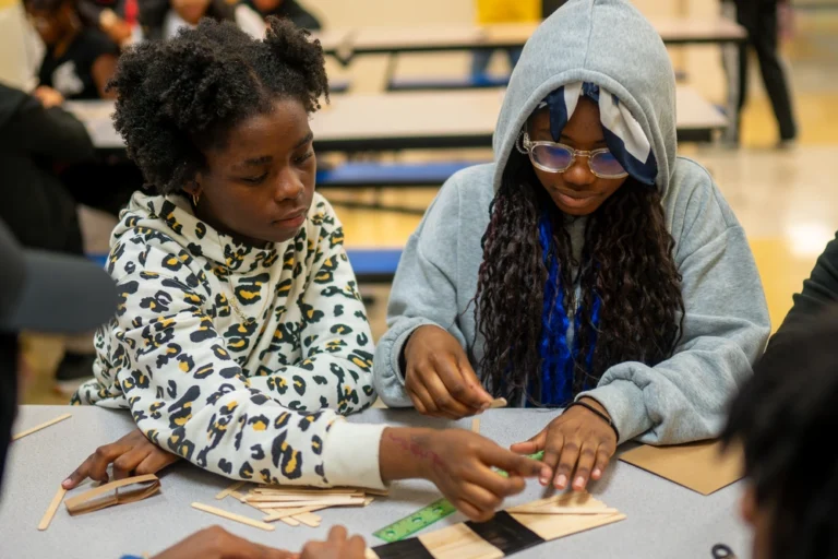 Two girls work together on a craft project with popsicle sticks at a table in a classroom or activity space.