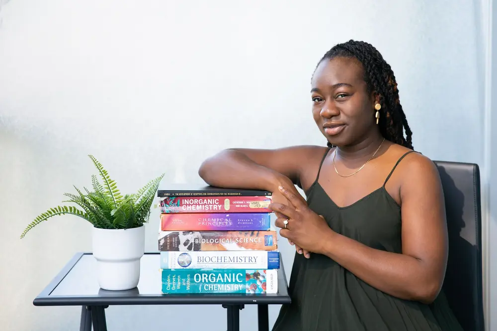 Woman in a black dress sits beside a stack of science textbooks and a potted plant on a small table.