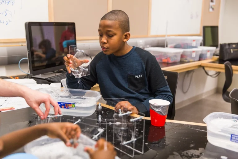 A boy in a classroom blows bubbles using a plastic frame and container of soapy water.