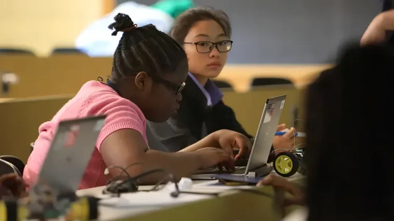 Two students sitting at desks using laptops, one typing and the other looking up attentively in a classroom.