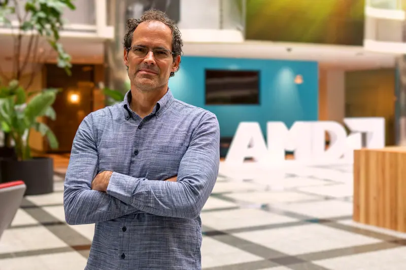 Man in a blue shirt stands with arms crossed in a modern lobby with large AMD letters in the background.
