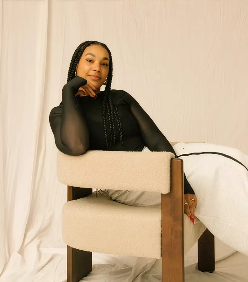Woman with long braids in a black top sits sideways on a beige chair, resting her head on her hand.