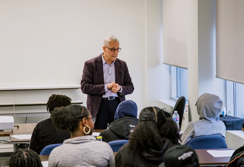 A man stands and speaks to a group of students seated in a classroom.