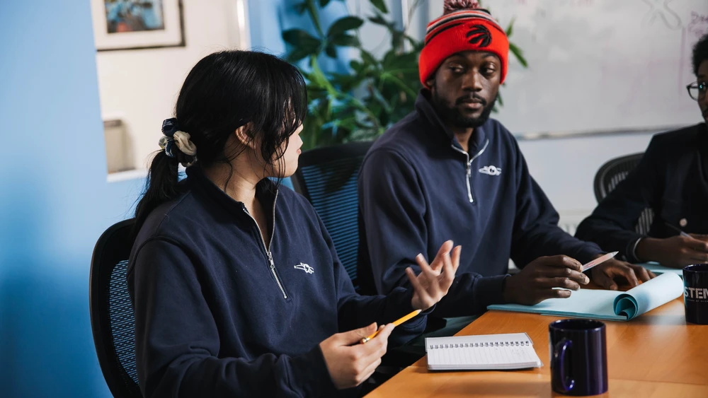 Two people in navy sweaters talk at a meeting table with notebooks, mugs, and papers in front of them.