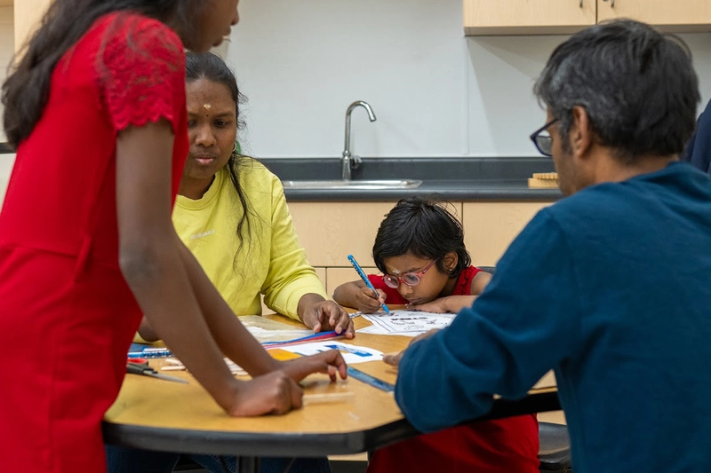 A family sits around a table, helping a young child with drawing and homework activities.