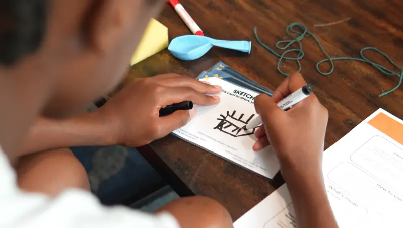 Child drawing a sketch with a black marker on a card at a table with papers, string, balloon, and marker.