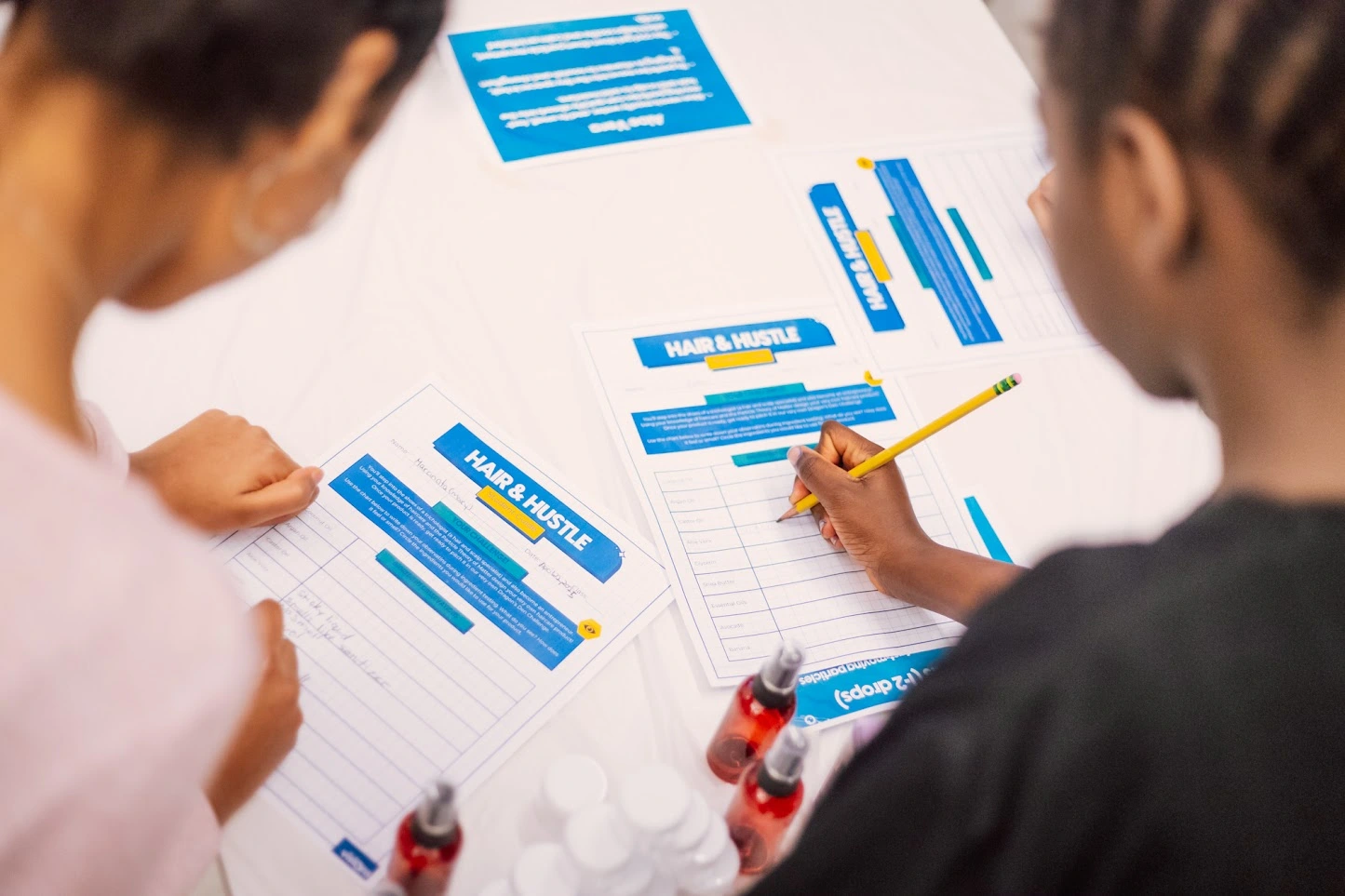 Two people fill out HAIR & HUSTLE forms at a table with pens, sanitizer bottles, and blue information sheets.