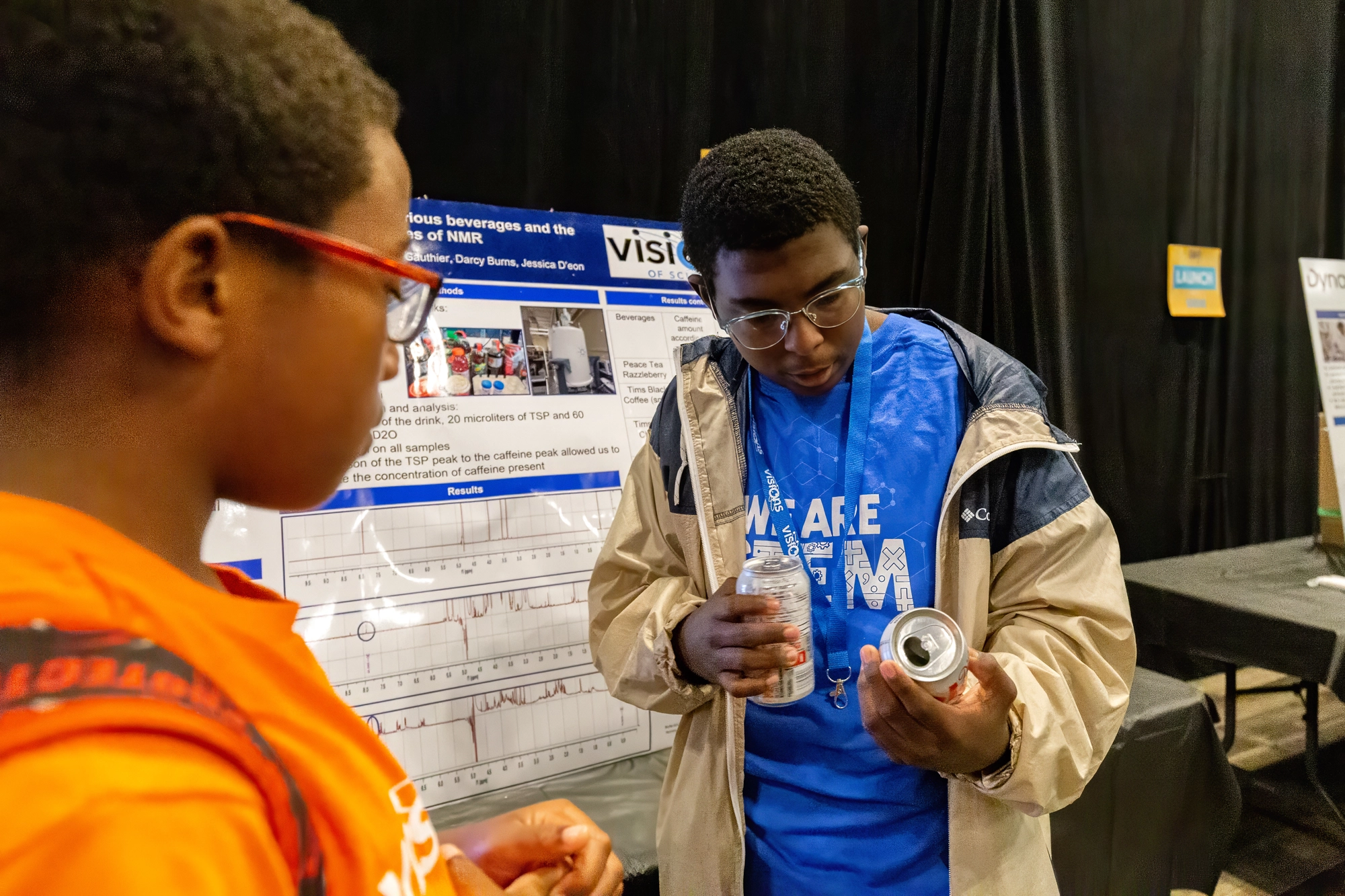 Two students examine a can in front of a science fair poster display at an event.