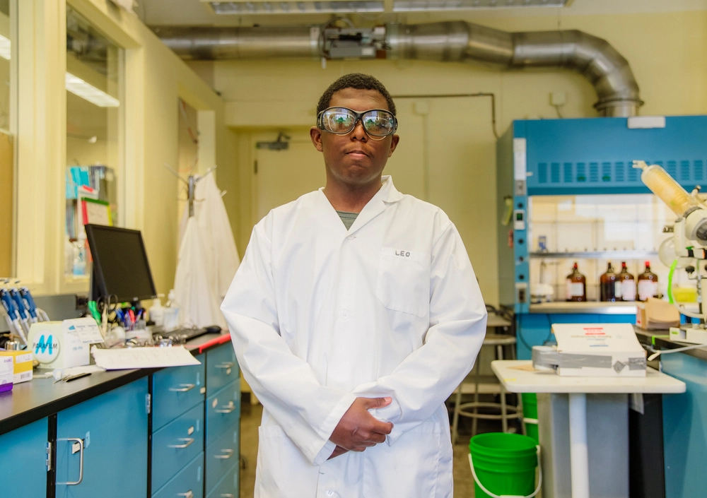 A person in a white lab coat and safety glasses stands in a laboratory with blue cabinets and equipment.
