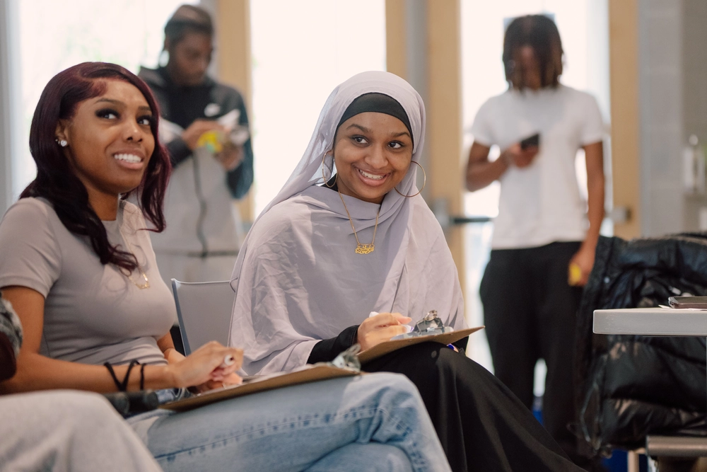Two young women smiling and writing on clipboards, with two people standing in the background.