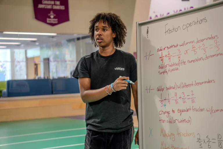 A person explains fraction operations on a whiteboard in a classroom or gym setting.