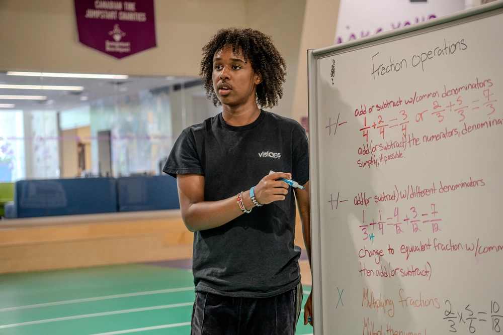 A person explains fraction operations on a whiteboard in a classroom or gym setting.