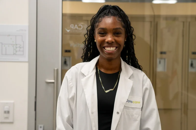 A woman in a white lab coat smiles, standing indoors in a professional or medical setting.