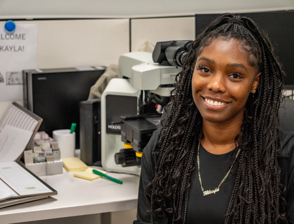 A woman with long braids smiles at a desk with a microscope and lab equipment in an office.