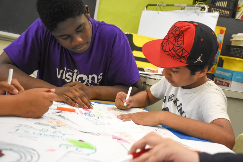 Two boys drawing together at a table in a classroom, focused on colorful artwork.