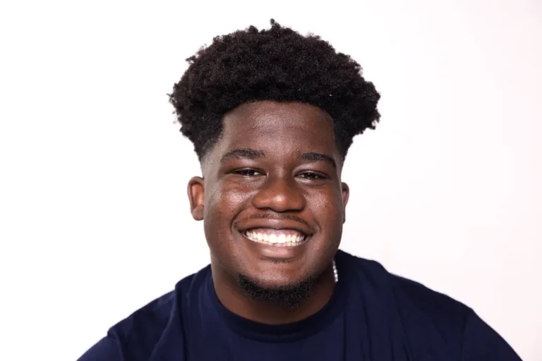 A young man with curly hair and a dark shirt smiles broadly against a plain white background.