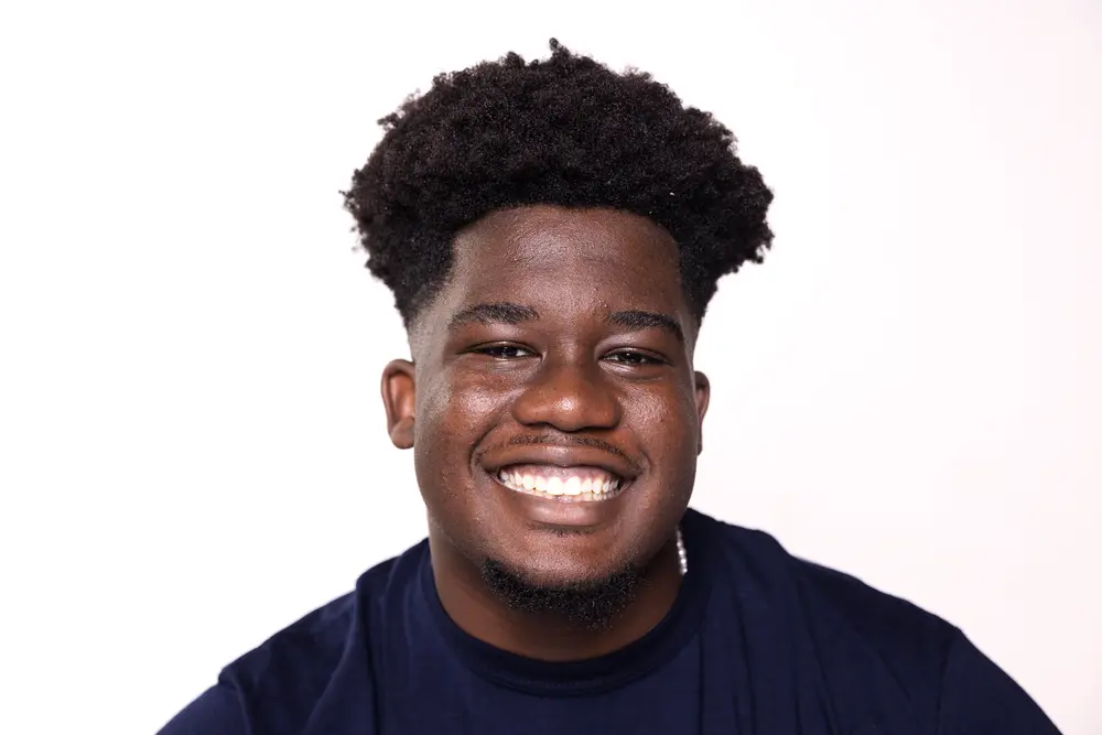 A young man with curly hair and a dark shirt smiles broadly against a plain white background.