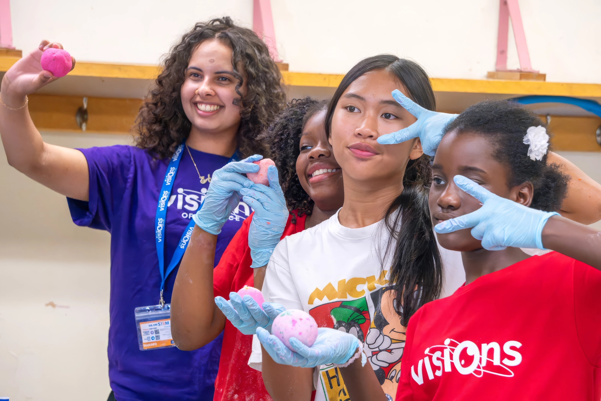 Four girls, some wearing gloves, smile and pose with colorful balls in a classroom setting.
