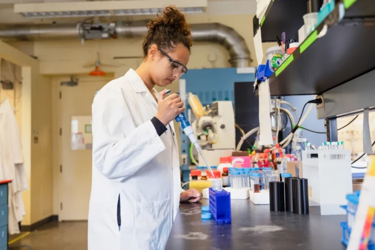 A scientist in a lab coat uses a pipette at a laboratory workbench with equipment and test tubes.