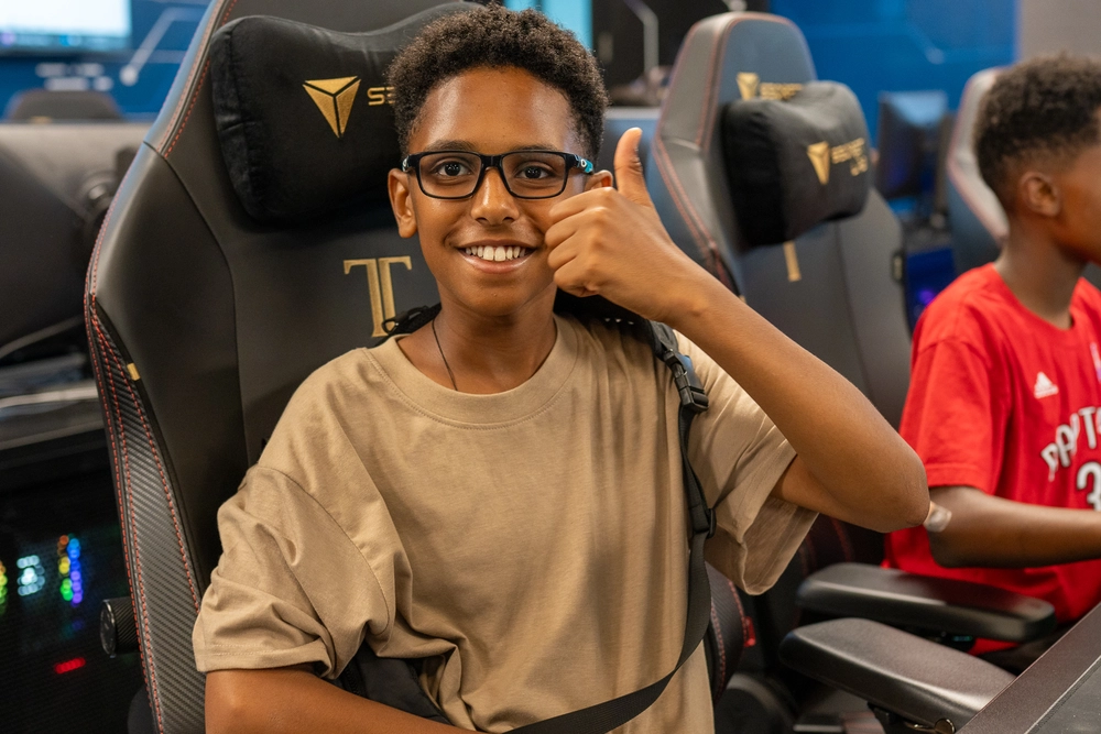 Smiling boy with glasses gives a thumbs up while sitting in a gaming chair at an esports venue.