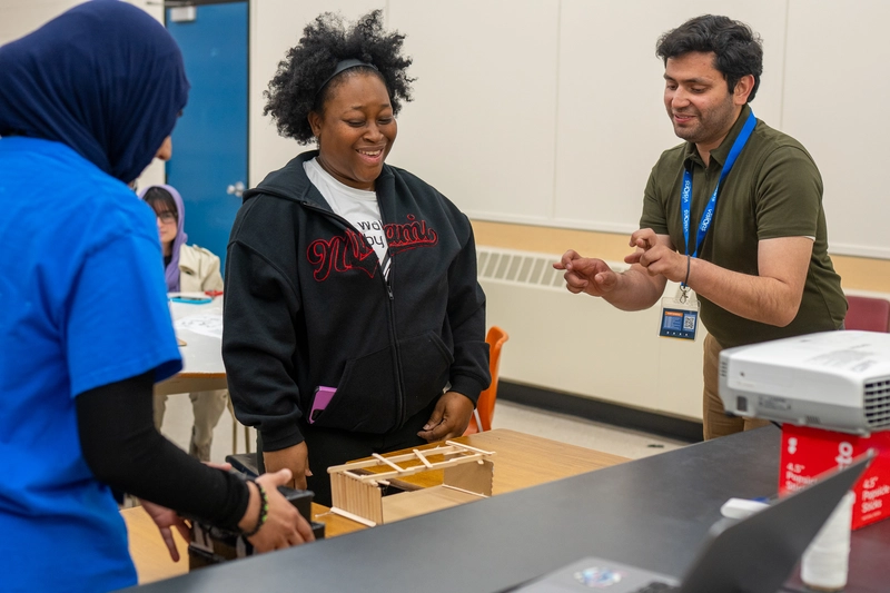 Three people smile and interact around a small wooden structure on a classroom table.
